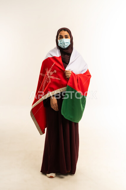 الوقاية من الامراض واتباع الاجراءات الاحترازية، إحياء ذكرى تأسيس الدولة 1775 م، حب الوطن والانتماء إليه، بورتريه لإمرأة عربية خليجية عمانية ترتدي الحجاب والعباءة والكمامة تضع علم البلد على كتفها، ذكرى اليوم الوطني العماني 18 نوفمبر، الجسم بالطول الكامل، خلفية بيضاء
