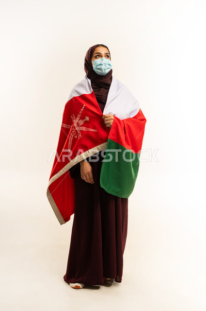 الوقاية من الامراض واتباع الاجراءات الاحترازية، إحياء ذكرى تأسيس الدولة 1775 م، حب الوطن والانتماء إليه، بورتريه لإمرأة عربية خليجية عمانية ترتدي الحجاب والعباءة والكمامة تضع علم البلد على كتفها، ذكرى اليوم الوطني العماني 18 نوفمبر، الجسم بالطول الكامل، خلفية بيضاء