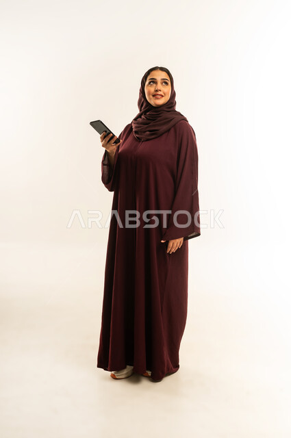 الدردشة مع الأهل والأصدقاء، إيماءات السعادة والسرور، استخدام الاجهزة التقنية الحديثة، بورتريه لامرأة عربية خليجية عمانية محجبة تحمل بيدها الهاتف المحمول، شابة سعودية تقوم بتصفح وسائل التواصل الاجتماعي، الجسم بالطول الكامل، خلفية بيضاء