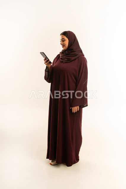 إيماءات السعادة والسرور، الدردشة مع الأهل والأصدقاء، استخدام الاجهزة التقنية الحديثة، بورتريه لامرأة عربية خليجية عمانية محجبة تحمل بيدها الهاتف المحمول، شابة سعودية تقوم بتصفح وسائل التواصل الاجتماعي، الجسم بالطول الكامل، خلفية بيضاء