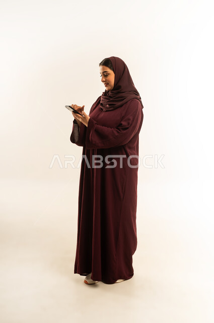 إيماءات السعادة والسرور، الدردشة مع الأهل والأصدقاء، استخدام الاجهزة التقنية الحديثة، بورتريه لامرأة عربية خليجية عمانية محجبة تحمل بيدها الهاتف المحمول، شابة سعودية تقوم بتصفح وسائل التواصل الاجتماعي، الجسم بالطول الكامل، خلفية بيضاء
