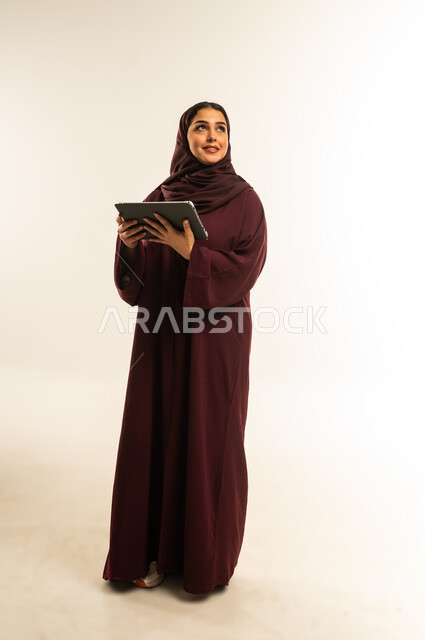 استخدام جهاز تقني حديث ومتطور، بورتريه لامرأة عربية خليجية عمانية ترتدي العباءة والحجاب تستخدم الجهاز اللوحي، شابة سعودية تقوم بإنجاز المهام المطلوبة عن بعد عبر التابلت، دمج التكنولوجيا بالعمل، الجسم بالطول الكامل، خلفية بيضاء