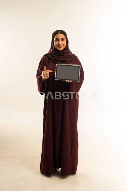 عرض شاشة سوداء فارغة، دمج التكنولوجيا بالعمل، استخدام جهاز تقني حديث ومتطور، بورتريه لامرأة عربية خليجية عمانية ترتدي العباءة والحجاب تستخدم الجهاز اللوحي، شابة سعودية تقوم بإنجاز المهام المطلوبة عن بعد عبر التابلت، الجسم بالطول الكامل، خلفية بيضاء
