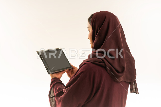 دمج التكنولوجيا بالعمل، استخدام جهاز تقني حديث ومتطور، بورتريه مقرب من الخلف لامرأة عربية خليجية عمانية ترتدي العباءة والحجاب تستخدم الجهاز اللوحي، شابة سعودية تقوم بإنجاز المهام المطلوبة عن بعد عبر التابلت، خلفية بيضاء
