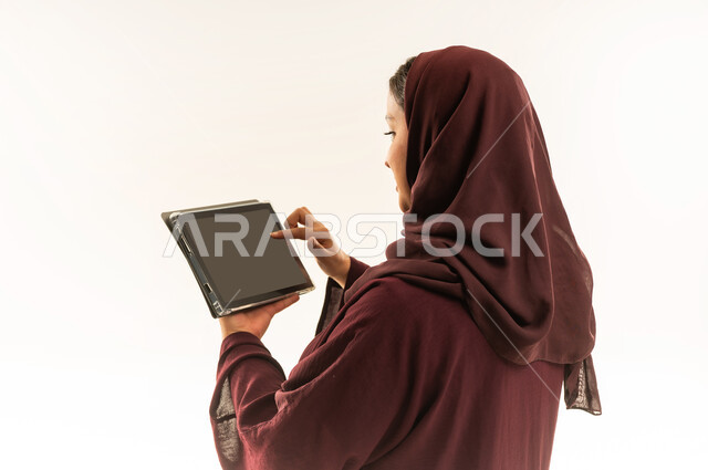دمج التكنولوجيا بالعمل، استخدام جهاز تقني حديث ومتطور، بورتريه مقرب من الخلف لامرأة عربية خليجية عمانية ترتدي العباءة والحجاب تستخدم الجهاز اللوحي، شابة سعودية تقوم بإنجاز المهام المطلوبة عن بعد عبر التابلت، خلفية بيضاء