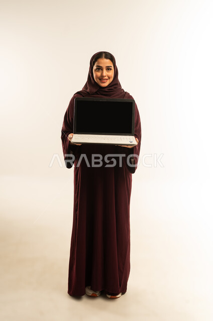 Integrating technology into work, a young Saudi woman displays a blank black screen on her laptop, completing tasks remotely, using a modern and advanced technical device, portrait of a smiling Omani Gulf Arab woman wearing an abaya and hijab using a laptop, full body, white background