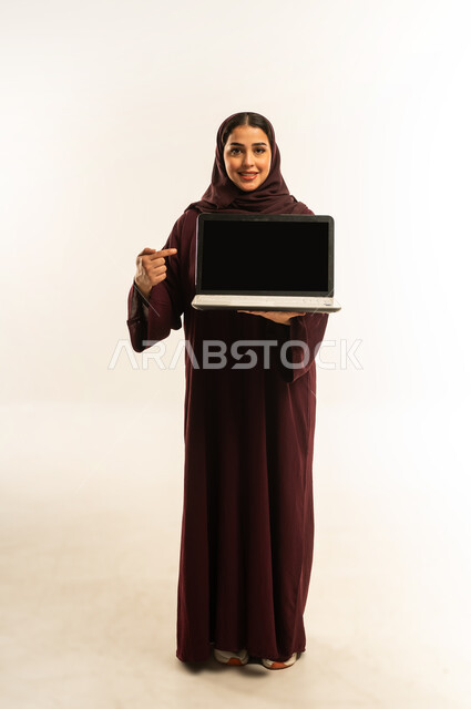 Integrating technology into work, a young Saudi woman displays a blank black screen on her laptop, completing tasks remotely, using a modern and advanced technical device, portrait of a smiling Omani Gulf Arab woman wearing an abaya and hijab using a laptop, full body, white background