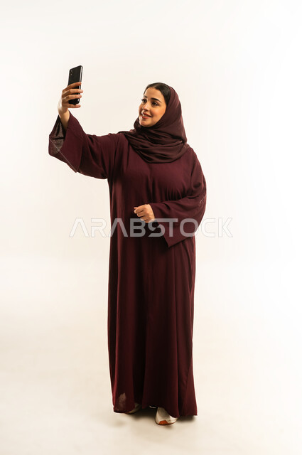 استخدام الاجهزة التقنية الحديثة، الدردشة مع الأهل والأصدقاء، إيماءات السعادة والسرور، بورتريه لامرأة عربية خليجية عمانية محجبة تحمل بيدها الهاتف المحمول، شابة سعودية تقوم بتصفح وسائل التواصل الاجتماعي، الجسم بالطول الكامل، خلفية بيضاء