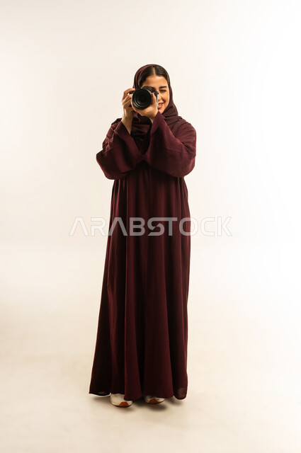 مهنة ووظيفة نسائية, مصورة سعودية تمارس هواية التصوير، مفهوم التصوير الفوتوغرافي باستخدام كاميرات رقمية عالية الدقة, بورتريه لإمراة عربية خليجية عمانية ترتدي الحجاب والعباءة تقوم بالتقاط صورة تذكارية عبر كاميرا فوتوغرافية, الجسم بالطول الكامل، خلفية بيضاء