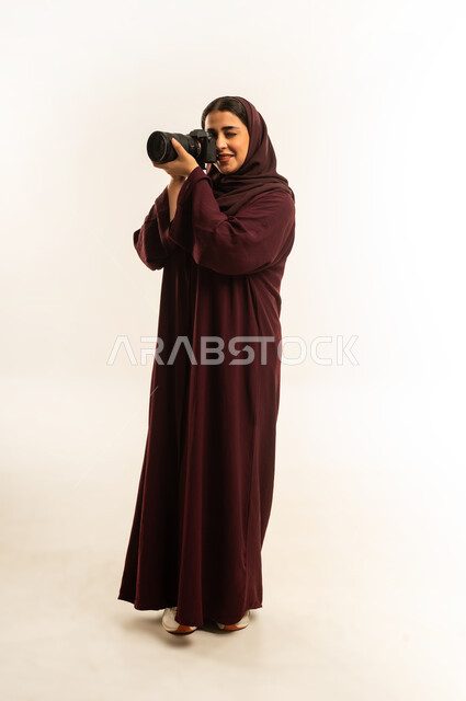 مصورة سعودية تمارس هواية التصوير، مهنة ووظيفة نسائية, مفهوم التصوير الفوتوغرافي باستخدام كاميرات رقمية عالية الدقة, بورتريه لإمراة عربية خليجية عمانية ترتدي الحجاب والعباءة تقوم بالتقاط صورة تذكارية عبر كاميرا فوتوغرافية, الجسم بالطول الكامل، خلفية بيضاء