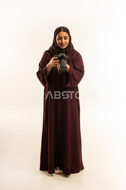 مهنة ووظيفة نسائية, مصورة سعودية تمارس هواية التصوير، مفهوم التصوير الفوتوغرافي باستخدام كاميرات رقمية عالية الدقة, بورتريه لإمراة عربية خليجية عمانية ترتدي الحجاب والعباءة تقوم بالتقاط صورة تذكارية عبر كاميرا فوتوغرافية, الجسم بالطول الكامل، خلفية بيضاء