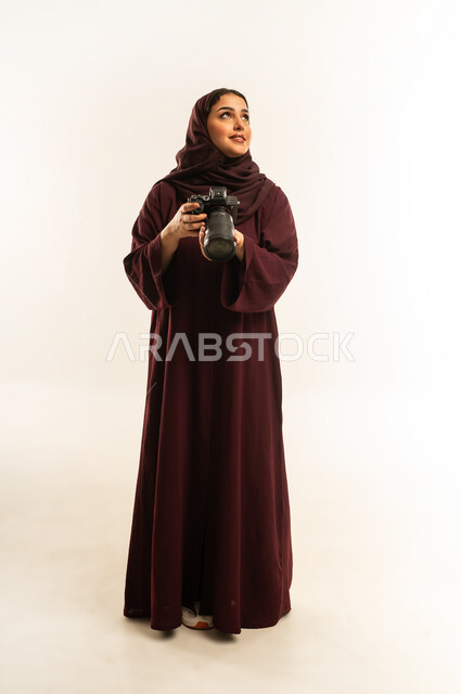 مصورة سعودية تمارس هواية التصوير، مهنة ووظيفة نسائية, مفهوم التصوير الفوتوغرافي باستخدام كاميرات رقمية عالية الدقة, بورتريه لإمراة عربية خليجية عمانية ترتدي الحجاب والعباءة تقوم بالتقاط صورة تذكارية عبر كاميرا فوتوغرافية, الجسم بالطول الكامل، خلفية بيضاء
