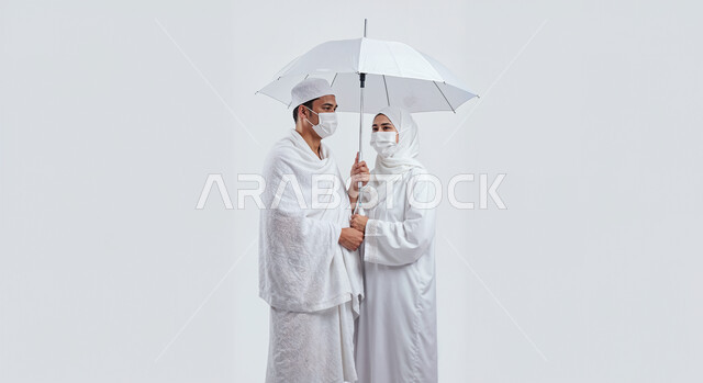 التظلل من اشعة الشمس باستخدام المظلة، مفهوم أركان الإسلام وأداء الفرائض الدينية، بورتريه مقرب لرجل حاج خليجي عربي سعودي يرتدي ثياب الإحرام يقف بجانب زوجته باستقامة، تأدية مناسك الحج والعمرة، العبادة والتقرب إلى الله، خلفية بيضاء
