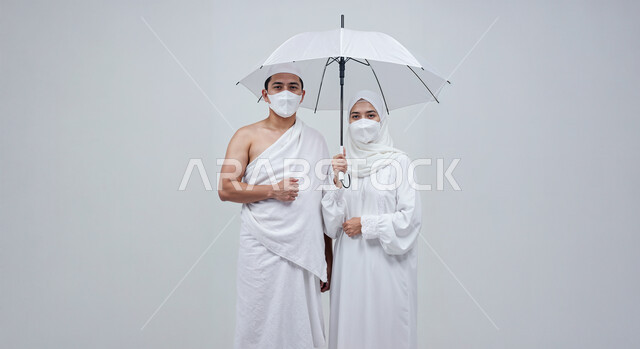 التظلل من اشعة الشمس باستخدام المظلة، مفهوم أركان الإسلام وأداء الفرائض الدينية، بورتريه مقرب لرجل حاج خليجي عربي سعودي يرتدي ثياب الإحرام يقف بجانب زوجته باستقامة، تأدية مناسك الحج والعمرة، العبادة والتقرب إلى الله، خلفية بيضاء