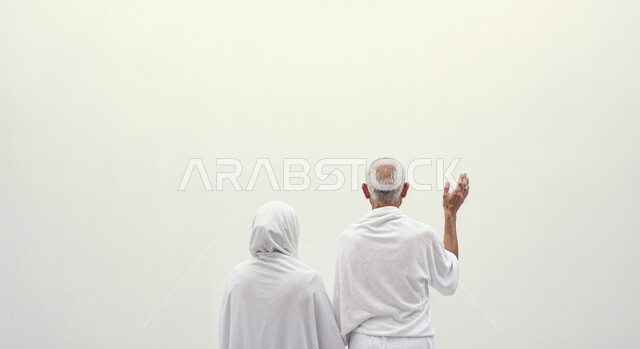 رفع اليدين والدعاء بخشوع وتضرع، مفهوم أركان الإسلام وأداء الفرائض الدينية، بورتريه من الخلف لشاب حاج خليجي عربي سعودي يرتدي ثياب الإحرام يقف بجانب زوجته باستقامة، تأدية مناسك الحج والعمرة، العبادة والتقرب إلى الله، خلفية بيضاء