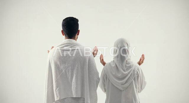 رفع اليدين والدعاء بخشوع وتضرع، مفهوم أركان الإسلام وأداء الفرائض الدينية، بورتريه من الخلف لشاب حاج خليجي عربي سعودي يرتدي ثياب الإحرام يقف بجانب زوجته باستقامة، تأدية مناسك الحج والعمرة، العبادة والتقرب إلى الله، خلفية بيضاء
