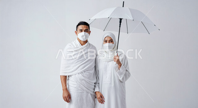 التظلل من اشعة الشمس باستخدام المظلة، مفهوم أركان الإسلام وأداء الفرائض الدينية، بورتريه مقرب لرجل حاج خليجي عربي سعودي يرتدي ثياب الإحرام يقف بجانب زوجته باستقامة، تأدية مناسك الحج والعمرة، العبادة والتقرب إلى الله، خلفية بيضاء