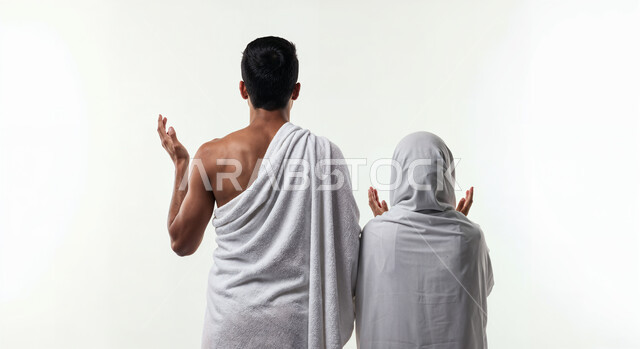 رفع اليدين والدعاء بخشوع وتضرع، مفهوم أركان الإسلام وأداء الفرائض الدينية، بورتريه من الخلف لشاب حاج خليجي عربي سعودي يرتدي ثياب الإحرام يقف بجانب زوجته باستقامة، تأدية مناسك الحج والعمرة، العبادة والتقرب إلى الله، خلفية بيضاء