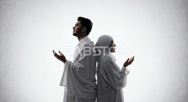 رفع اليدين والدعاء بخشوع وتضرع، مفهوم أركان الإسلام وأداء الفرائض الدينية، بورتريه مقرب لشاب حاج خليجي عربي سعودي يرتدي ثياب الإحرام يقف بجانب زوجته باستقامة بتعابير السرور، تأدية مناسك الحج والعمرة، العبادة والتقرب إلى الله، خلفية بيضاء