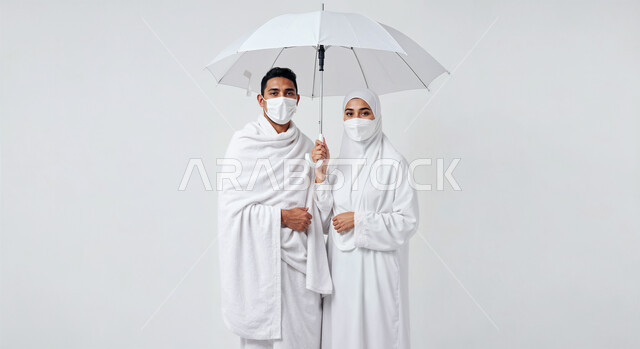 التظلل من اشعة الشمس باستخدام المظلة، مفهوم أركان الإسلام وأداء الفرائض الدينية، بورتريه مقرب لرجل حاج خليجي عربي سعودي يرتدي ثياب الإحرام يقف بجانب زوجته باستقامة، تأدية مناسك الحج والعمرة، العبادة والتقرب إلى الله، خلفية بيضاء
