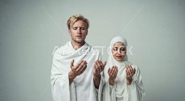 رفع اليدين والدعاء بخشوع وتضرع، مفهوم أركان الإسلام وأداء الفرائض الدينية، بورتريه مقرب لشاب حاج خليجي عربي سعودي يرتدي ثياب الإحرام يقف بجانب زوجته باستقامة بتعابير السرور، تأدية مناسك الحج والعمرة، العبادة والتقرب إلى الله، خلفية بيضاء