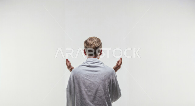 مفهوم أركان الإسلام وأداء الفرائض الدينية، رفع اليدين والدعاء بخشوع وتضرع، بورتريه مقرب من الخلف لحاج خليجي عربي سعودي يرتدي ثياب الإحرام يقوم بتأدية مناسك الحج والعمرة، العبادة والتقرب إلى الله، خلفية بيضاء