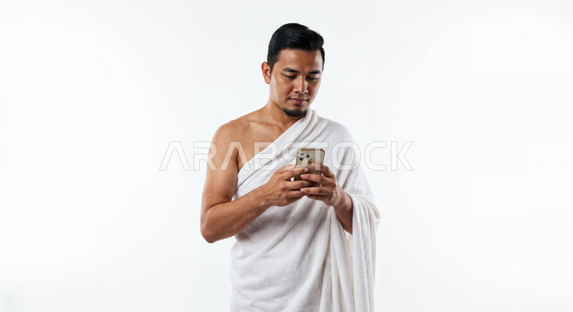 Using the Hajj guide application on mobile, performing Hajj and Umrah rituals, close-up portrait of a Saudi Arabian Gulf Muslim pilgrim wearing Ihram clothes using a mobile phone, white background