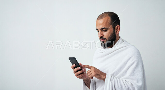 Using the Hajj guide application on mobile, performing Hajj and Umrah rituals, close-up portrait of a Saudi Arabian Gulf Muslim pilgrim wearing Ihram clothes using a mobile phone, white background