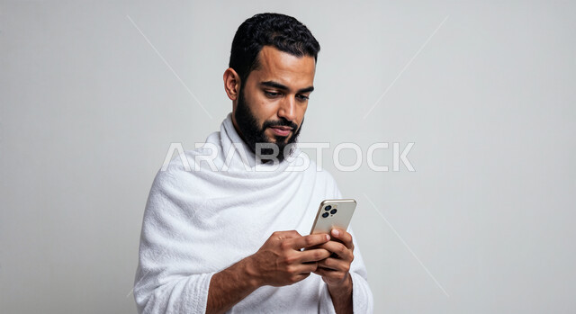 Using the Hajj guide application on mobile, performing Hajj and Umrah rituals, close-up portrait of a Saudi Arabian Gulf Muslim pilgrim wearing Ihram clothes using a mobile phone, white background