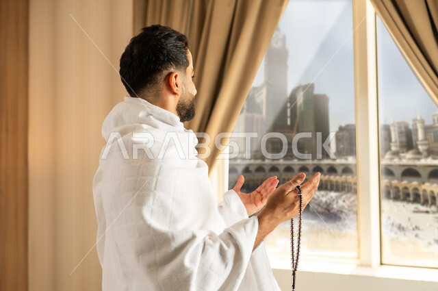العبادة والتقرب إلى الله تعالى، الاطلالة على ساحة الحرم المكي الشريف، صورة من الجانب لرجل يرتدي ثياب الاحرام ينظر الى الكعبة المشرفة ويرفع يديه بتعابير الدعاء والتضرع, أداء مناسك الحج والعمرة، مكان ومعلم اسلامي ديني مقدس في المملكة العربية السعودية