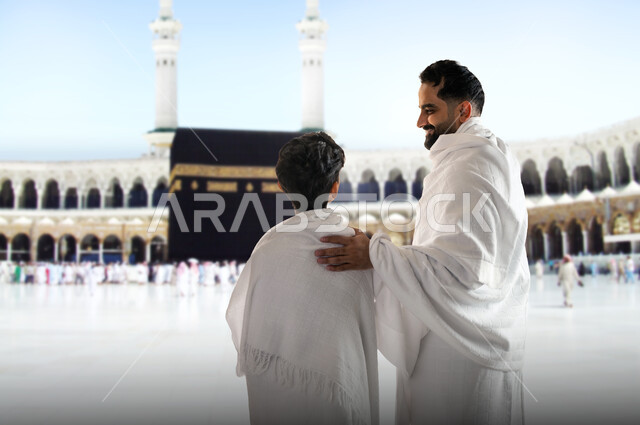 Standing in the courtyard of the Grand Mosque in Mecca, a picture from the back of a man wearing Ihram clothes contemplating the Holy Kaaba in Mecca with his son, the destination of Muslims from all over the world, performing the Hajj and Umrah rituals, a sacred Islamic religious place and landmark in the Kingdom of Saudi Arabia, worship and drawing closer to God Almighty