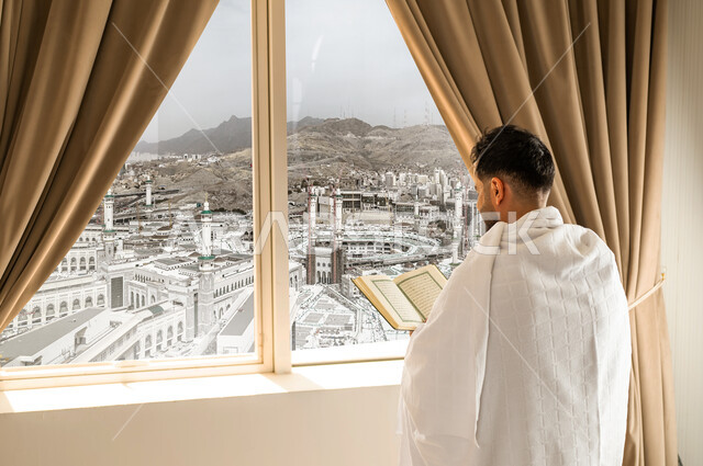 قراءة وتلاوة القرآن الكريم، العبادة والتقرب إلى الله تعالى، الاطلالة على ساحة الحرم المكي الشريف، صورة من الجانب لرجل يرتدي ثياب الاحرام ينظر الى الكعبة المشرفة يمسك بيده المصحف الشريف, أداء مناسك الحج والعمرة، مكان ومعلم اسلامي ديني مقدس في المملكة العربية السعودية