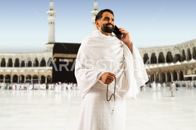 إجراء مكالمه هاتفيه عبر الجوال, رجل مسلم خليجي عربي سعودي يرتدي ملابس الاحرام  يقف في الحرم المكي الشريف ، أداء الصلاة ومناسك الحج والعمره في المسجد الحرام في مكة المكرمه، ، موسم الحج في شهر ذو الحجه