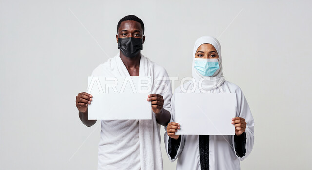 لوحة بيضاء فارغة، مفهوم أركان الإسلام وأداء الفرائض الدينية، بورتريه مقرب لرجل حاج يرتدي ثياب الإحرام والكمامة يقف بجانب زوجته باستقامة يمسكان موك اب ابيض، تأدية مناسك الحج والعمرة، العبادة والتقرب إلى الله، اتباع الاجراءات الوقائية من العدوى، خلفية بيضاء