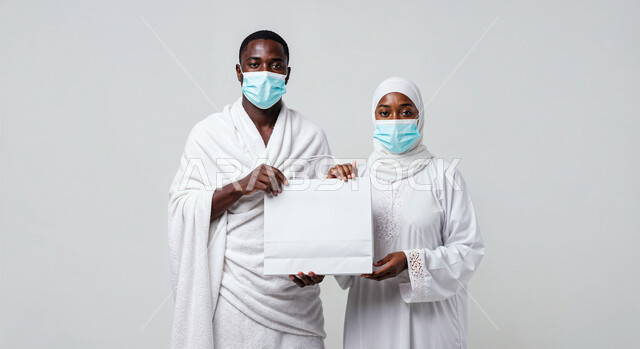 لوحة بيضاء فارغة، مفهوم أركان الإسلام وأداء الفرائض الدينية، بورتريه مقرب لرجل حاج يرتدي ثياب الإحرام والكمامة يقف بجانب زوجته باستقامة يمسكان موك اب ابيض، تأدية مناسك الحج والعمرة، العبادة والتقرب إلى الله، اتباع الاجراءات الوقائية من العدوى، خلفية بيضاء