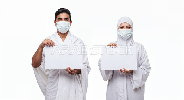 لوحة بيضاء فارغة، مفهوم أركان الإسلام وأداء الفرائض الدينية، بورتريه مقرب لرجل حاج يرتدي ثياب الإحرام والكمامة يقف بجانب زوجته باستقامة يمسكان موك اب ابيض، تأدية مناسك الحج والعمرة، العبادة والتقرب إلى الله، اتباع الاجراءات الوقائية من العدوى، خلفية بيضاء