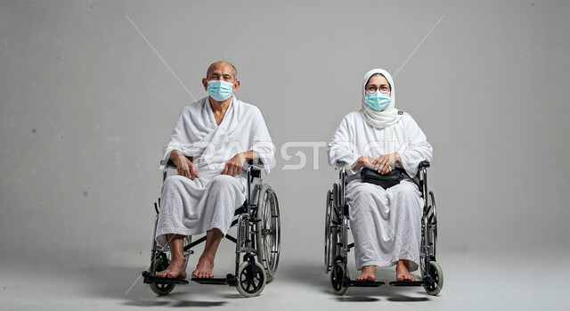 Following preventive measures against the Corona virus, the concept of the pillars of Islam and performing religious duties, a portrait of a pilgrim with special needs wearing ihram clothes and a mask sitting in a wheelchair with his wife performing the Hajj and Umrah rituals, worship and drawing closer to God, white background