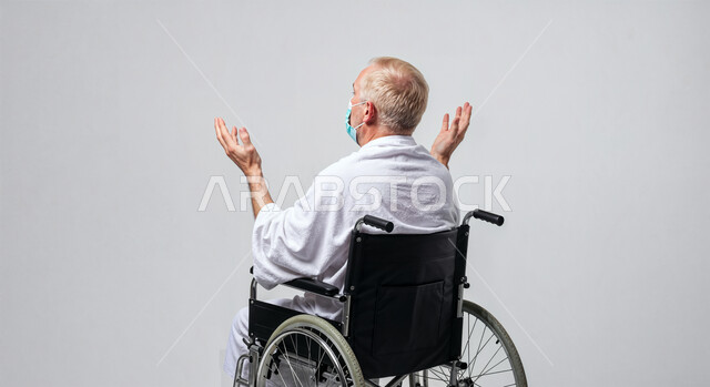 مفهوم أركان الإسلام وأداء الفريضة الدينية، رفع اليدين والدعاء بخشوع وتضرع، بورتريه لحاج من ذوي الهمم يرتدي ثياب الإحرام يجلس على كرسي متحرك يقوم بتأدية مناسك الحج والعمرة، العبادة والتقرب إلى الله، خلفية بيضاء