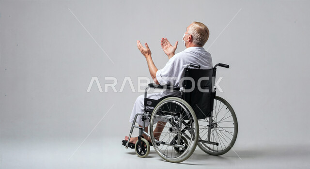 مفهوم أركان الإسلام وأداء الفريضة الدينية، رفع اليدين والدعاء بخشوع وتضرع، بورتريه لحاج من ذوي الهمم يرتدي ثياب الإحرام يجلس على كرسي متحرك يقوم بتأدية مناسك الحج والعمرة، العبادة والتقرب إلى الله، خلفية بيضاء