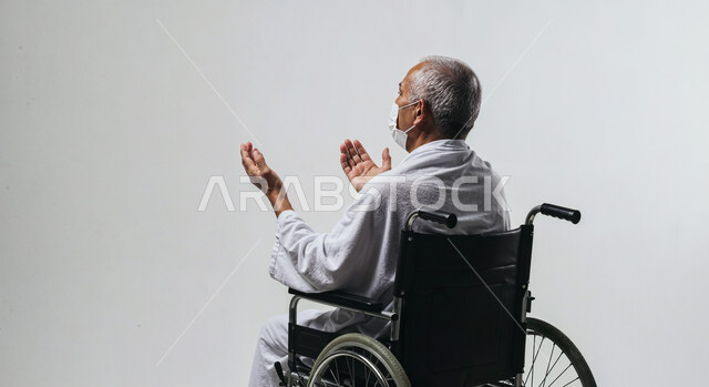 مفهوم أركان الإسلام وأداء الفريضة الدينية، رفع اليدين والدعاء بخشوع وتضرع، بورتريه لحاج من ذوي الهمم يرتدي ثياب الإحرام يجلس على كرسي متحرك يقوم بتأدية مناسك الحج والعمرة، العبادة والتقرب إلى الله، خلفية بيضاء