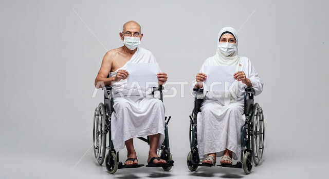 لوحة بيضاء فارغة، مفهوم أركان الإسلام وأداء الفرائض الدينية، بورتريه لرجل حاج من ذوي الهمم يرتدي ثياب الإحرام والكمامة يجلس على كرسي متحرك بجانب زوجته يمسكان موك اب ابيض، تأدية مناسك الحج والعمرة، العبادة والتقرب إلى الله، اتباع الاجراءات الوقائية، خلفية بيضاء