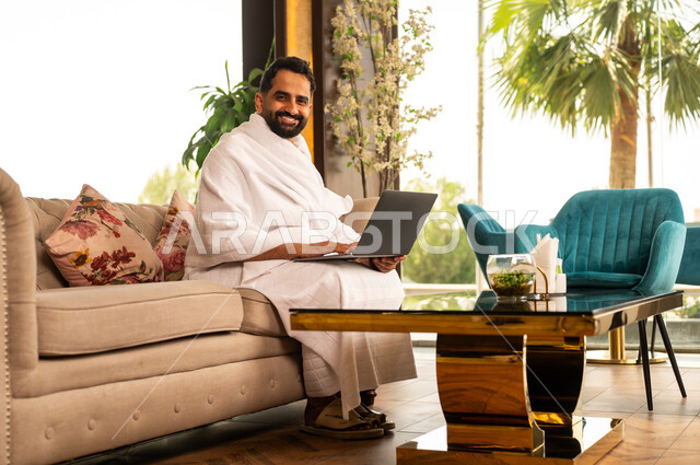 استخدام الأجهزة التقنية الحديثة في تصفح مواقع الارشادات لأداء مناسك الحج والعمرة، رجل مسلم خليجي عربي سعودي مبتسم يرتدي ملابس الاحرام يحمل بيده جهاز الحاسوب المحمول يجلس على كنبة مريحة في بهو الفندق، إيماءات السرور والسعادة