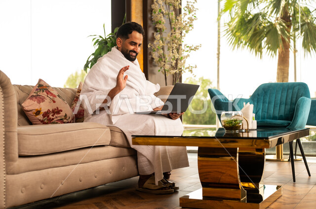 استخدام الأجهزة التقنية الحديثة في تصفح مواقع الارشادات لأداء مناسك الحج والعمرة، رجل مسلم خليجي عربي سعودي مبتسم يرتدي ملابس الاحرام يحمل بيده جهاز الحاسوب المحمول يجلس على كنبة مريحة في بهو الفندق، إيماءات السرور والسعادة