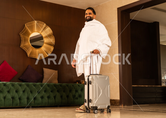 الاستعداد لأداء مناسك الحج والعمرة، العبادة والتقرب إلى الله تعالى، رجل عربي خليجي سعودي يرتدي ثياب الاحرام يمسك بيده حقيبة الامتعة يقف في بهو الفندق, زيارة مكان ومعلم اسلامي ديني مقدس في المملكة العربية السعودية