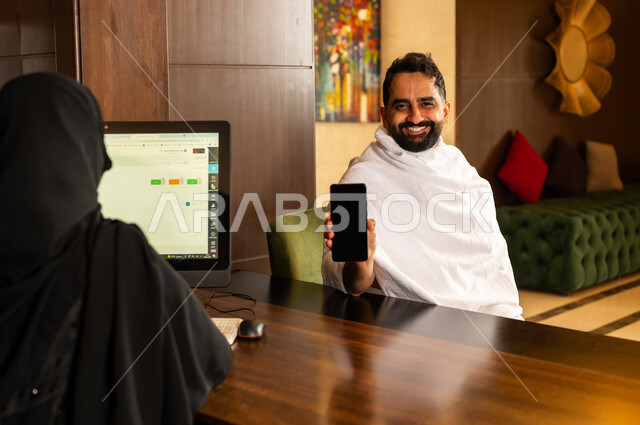 الدفع عن طريق بطاقة الائتمان البنكية عبر الهاتف المحمول, القيام بإجراءات الحجز والدخول لاتمام مناسك الحج والعمرة, منتجعات وفنادق مكة المكرمة في المملكة العربية السعودية، رجل عربي خليجي سعودي يرتدي لباس الاحرام يقوم بالدفع في مكتب الاستقبال للموظفة