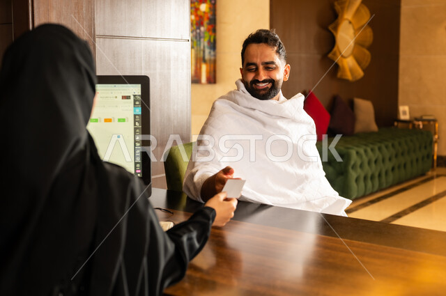 خدمة العملاء والاستقبال للحجاج والمعتمرين، نظام المفتاح الالكتروني لباب الغرفة، رجل مسلم عربي خليجي سعودي يرتدي لباس الاحرام يجلس أمام مكتب الإستقبال يقوم بأخذ البطاقة من الموظفة, فنادق ومنتجعات مكة المكرمة في المملكة العربية السعودية، تعابير البشاشة والسرور