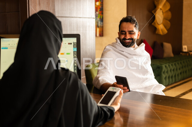 Payment by credit card via mobile phone, making reservations and entering to complete Hajj and Umrah rituals, Makkah Al-Mukarramah resorts and hotels in the Kingdom of Saudi Arabia, a Saudi Arabian Gulf man wearing Ihram clothing paying at the reception desk to the employee