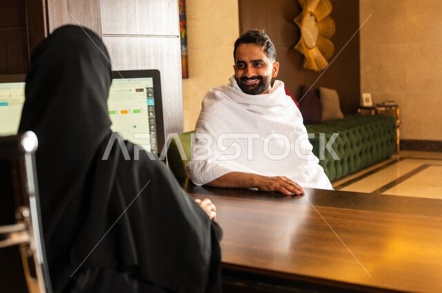 خدمة العملاء والاستقبال للحجاج والمعتمرين، رجل مسلم عربي خليجي سعودي يرتدي لباس الاحرام يجلس أمام مكتب الإستقبال للاستفسار عن أسعار الشقق السكنيه واتمام الحجز, فنادق ومنتجعات مكة المكرمة في المملكة العربية السعودية