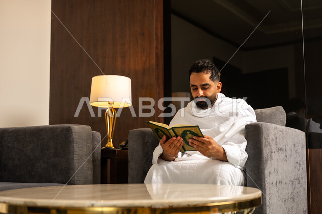 قراءة وتلاوة القرآن الكريم، العبادة والتقرب إلى الله تعالى، رجل عربي خليجي سعودي يرتدي ثياب الاحرام يمسك بيده المصحف الشريف يجلس على كنبة في بهو الفندق, أداء مناسك الحج والعمرة، مكان ومعلم اسلامي ديني مقدس في المملكة العربية السعودية