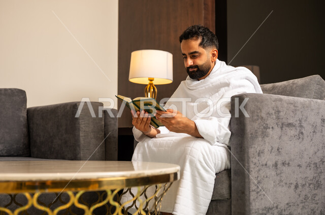 Reading and reciting the Holy Quran, worship and drawing closer to God Almighty, a Saudi Arabian Gulf man wearing Ihram clothes holding the Holy Quran in his hand sitting on a sofa in the hotel lobby, performing Hajj and Umrah rituals, a sacred Islamic religious place and landmark in the Kingdom of Saudi Arabia